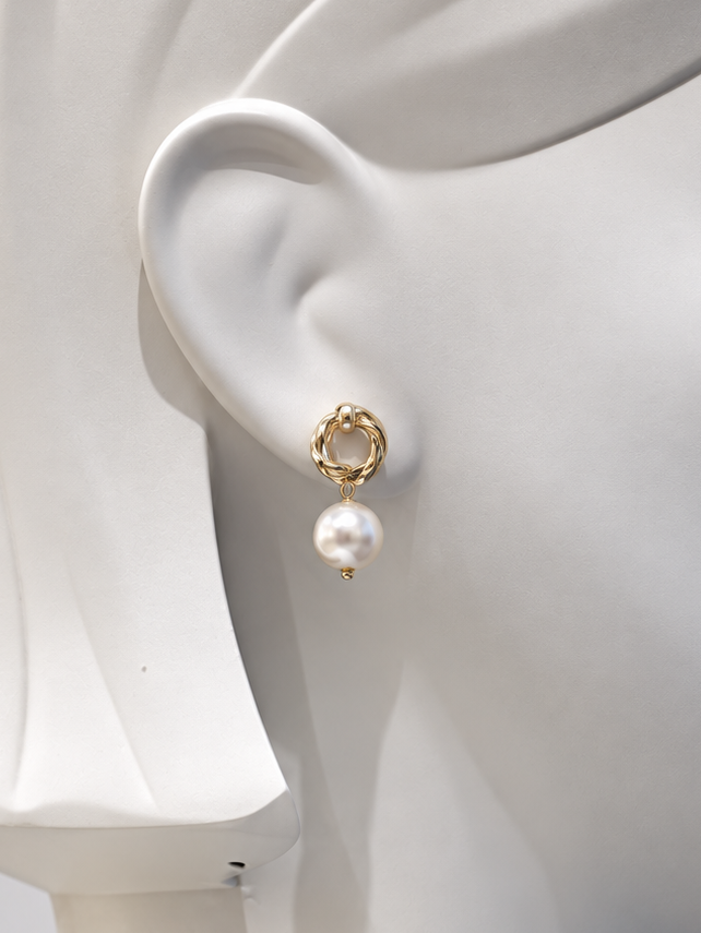 A close-up of a white mannequin head wearing a single gold hoop earring with a dangling white pearl, shown against a light gray background.